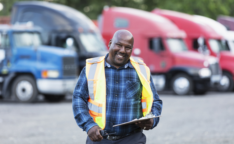 Portrait of a mature African-American man standing, holding a clipboard, with a fleet of semis parked in a row in the background. He is a truck driver, a manager, or perhaps the owner of this small freight transportation business. He is smiling at the camera.